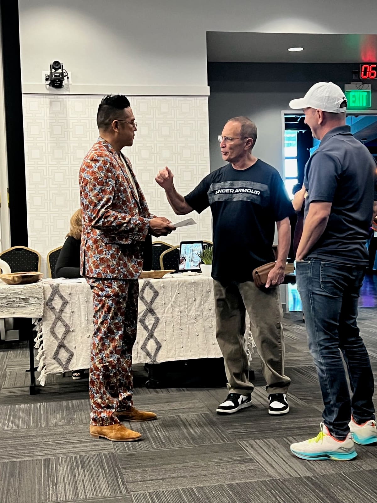 Man in patterned jacket speaking with two men at indoor event near table with tablet