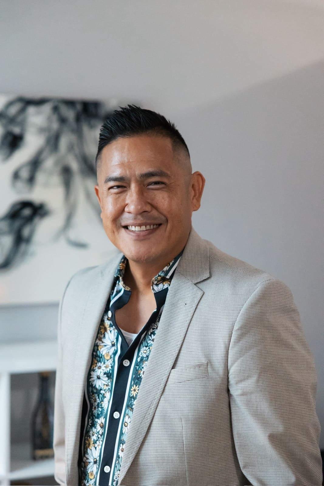 Professional headshot of a man wearing a light beige blazer and floral patterned shirt, smiling at the camera in a modern office setting