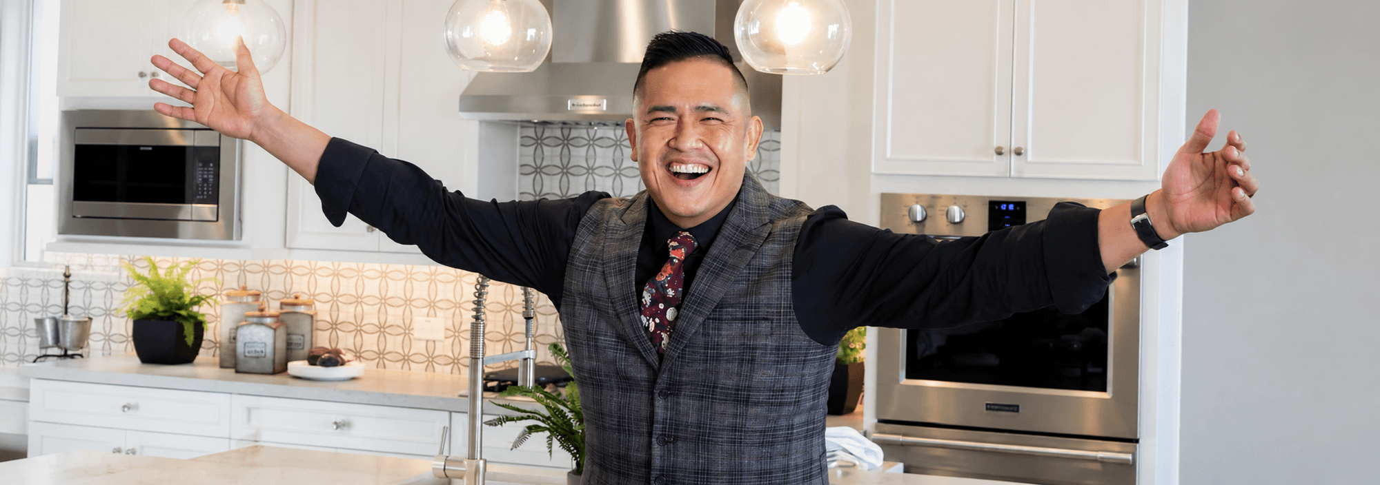 Man in blazer and patterned tie smiling with arms spread wide in modern kitchen